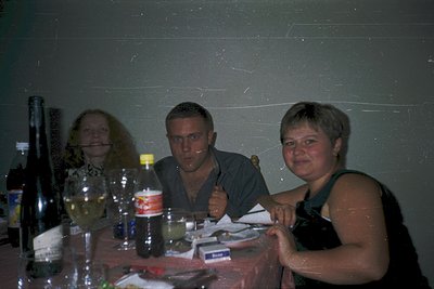 Indoor gathering with three adults seated at a table, likely a casual social event. The man in the center wears a dark top an...