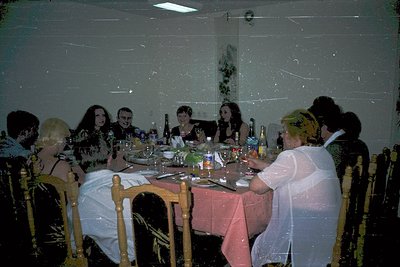 Vintage indoor gathering at a round table with 10 seated guests, dressed in formal attire (1970s-80s style). Wooden chairs, r...