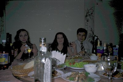 Three adults seated at a formal dinner table, mid-1990s Eastern Europe. Left: woman in dark blazer, holding wineglass; center...