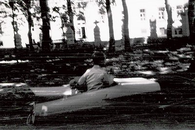 Black-and-white shot of a lone figure seated on a park bench, back to camera, in an urban park with blurred motion. Trees and...