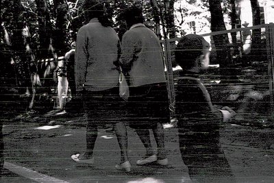 Blurred monochrome street scene featuring three figures in motion, likely mid-1970s urban setting. Two adults walk side-by-si...