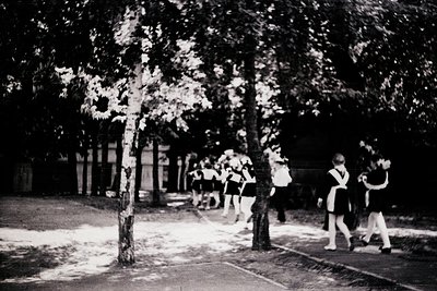 Black-and-white street scene of uniformed students walking in single file along a tree-lined path, likely a school or youth g...