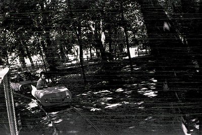 Vintage black-and-white shot of a vintage ice cream cart in a shaded urban park. The cart’s signage suggests a mid-20th-centu...
