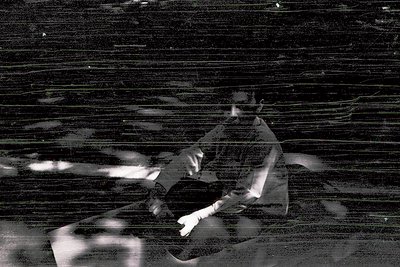 Vintage black-and-white photo of a seated figure against a textured wall, likely 1950s–1970s. The individual wears a light-co...