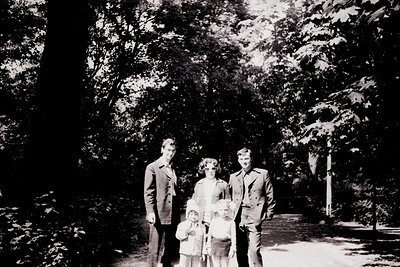 Family portrait in a wooded park setting, mid-20th century. Four adults and two children pose on a gravel path surrounded by ...
