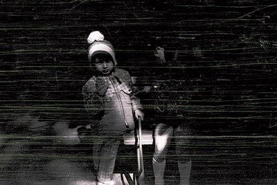 Vintage black-and-white photo of a child in winter attire—knit hat, coat, and mittens—holding a stick, blurred against a wood...