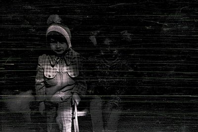 Black-and-white portrait of a child in mid-20th century attire—plaid shirt, knit cap, and suspenders—leaning on a cane agains...