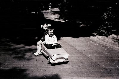 Two children in 1960s-style clothing play with a vintage toy car on a paved pathway. The girl in the foreground sits in the t...