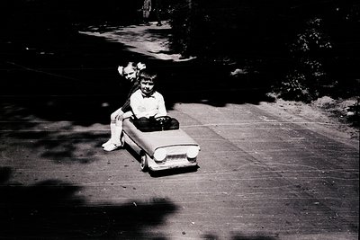 Vintage black-and-white photo of a child riding a toy car on a paved driveway, mid-20th century. The child wears a short-slee...