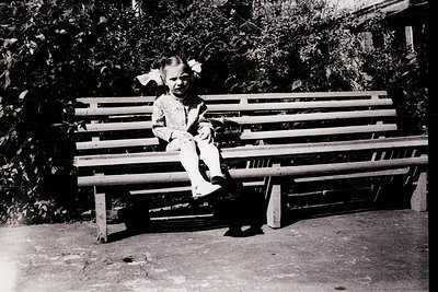 Young child in early 20th-century dress sits on a wooden bench, holding a small stuffed toy. Lush greenery and a brick pathwa...