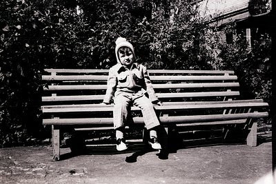 Mid-20th century black-and-white portrait of a child in winter attire—knit hat, coat, and boots—seated on a wooden park bench...