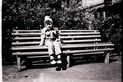 Black-and-white snapshot of a child in 1950s-60s attire—knit hat, long-sleeve shirt, and knee-length pants—sitting on a woode...