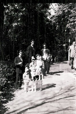 Family group photo in a wooded forest path, mid-20th century. Adults in suits and light jackets, children in dresses and shor...