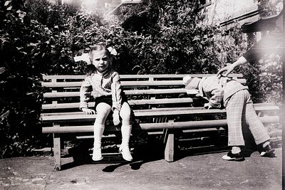 Vintage black-and-white photo of two children playing on a wooden bench in an outdoor setting, likely mid-20th century. Child...