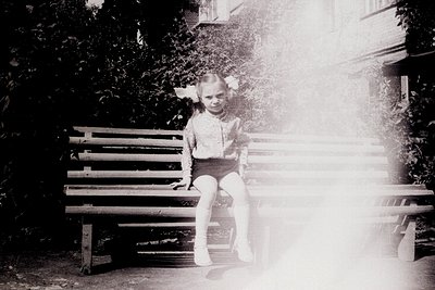Black-and-white vintage photo of a young girl sitting on a wooden bench in sunlight, backlit by light rays. She wears a short...