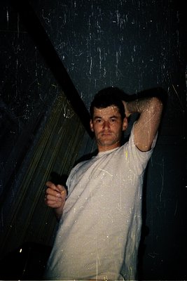 Portrait of a man in a dimly lit indoor setting, possibly a 1970s–1980s dance club or party. He stands with one hand on his h...