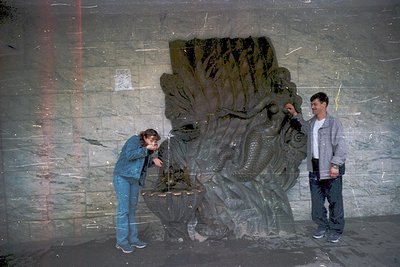 Two individuals inspecting a large, weathered concrete relief sculpture depicting a stylized fish or aquatic creature, likely...