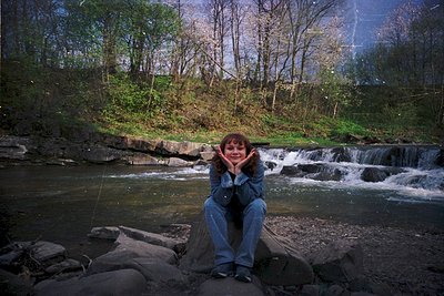 A person sits on a rock by a cascading stream, hands framing their face. Mid-20th century clothing (denim jacket, jeans) sugg...