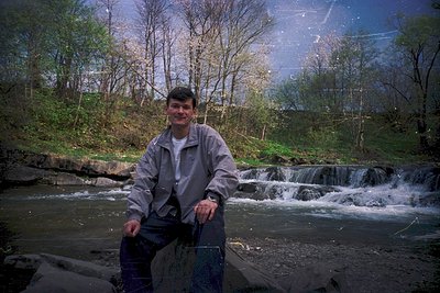 Man poses by a cascading river with a vintage fishing net, likely 1980s–1990s. Forest and rocky terrain in background. Casual...