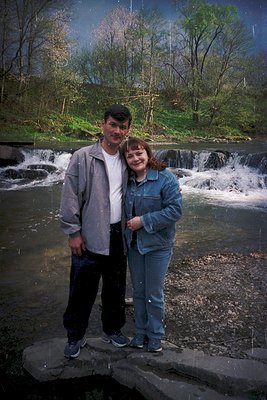 Couple posing by a cascading waterfall in a lush, wooded area, likely late 20th century. Man in a light gray jacket, woman in...