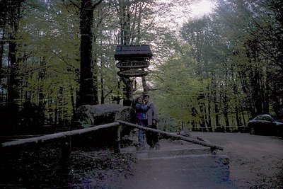 Vintage photo of a couple posing on a stone bridge under a wooden sign reading "Свещовски" (Sveshtarski) in a forested area. ...