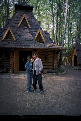 Log cabin with steep gabled roof and wooden trim, surrounded by lush forest. Two people pose in front, dressed in 1970s-style...