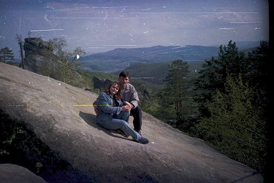 Vintage 1960s/70s couple posing on rocky cliff edge overlooking forested valley. Colorful, slightly faded film captures their...