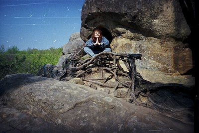 A person sits in a natural rock formation with twisted roots, framed by a cave-like opening under bright sunlight.