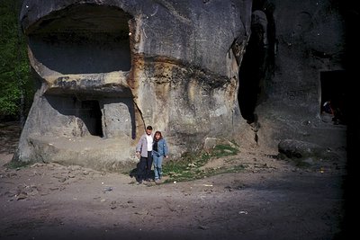 Two individuals pose in front of a massive concrete bunker carved into rock, likely from the Cold War era. The structure feat...