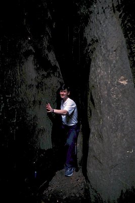 Indoor rock climbing scene with climber mid-route on a smooth, vertical limestone wall. The climber wears a white T-shirt, bl...