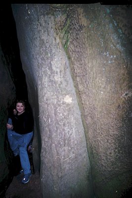 A person poses beside a massive, weathered stone column in a dimly lit underground chamber. The column features carved inscri...