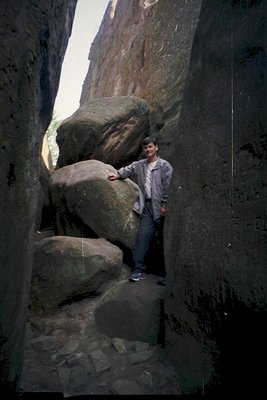 A person stands in a narrow, natural rock formation tunnel, leaning against a large boulder. The rugged, vertical rock walls ...
