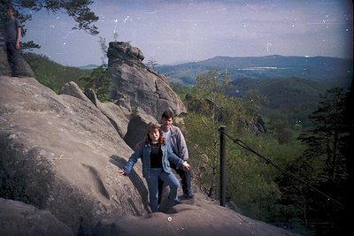 Two individuals pose on a rocky outcrop overlooking a forested valley, framed by vintage Polaroid-style borders. Clothing sug...