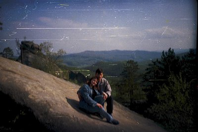 Vintage Polaroid-style shot of two individuals seated on rocky terrain, overlooking forested hills under a hazy sky. Distress...