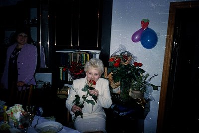 Vintage indoor celebration featuring an elderly woman in a white blouse holding a red rose, surrounded by bookshelves and flo...