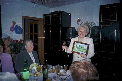 Vintage indoor celebration featuring a woman in 1970s formal wear holding a framed landscape painting, surrounded by older ad...