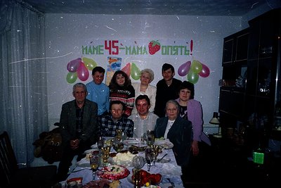 Family celebration for a 45th birthday in a modest indoor setting, likely Eastern Europe, 1980s–1990s. Nine adults pose aroun...