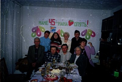 Family celebration for a 45th birthday in a modest indoor setting, likely late 20th century. Nine adults pose around a table ...