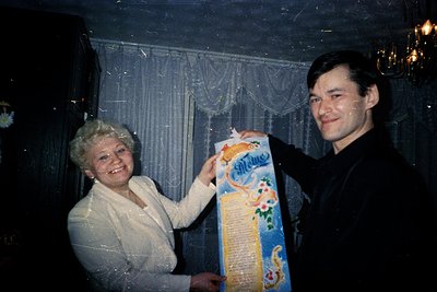 Vintage indoor portrait featuring a man and woman holding a rolled, illustrated scroll with Cyrillic text. The woman wears a ...