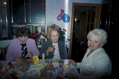 Three women in formal attire share a festive meal indoors, likely late 20th century. Center table displays vintage food conta...