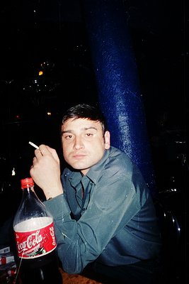Young man in a 1970s-style button-up shirt leans against a dark, reflective surface, holding a lit cigarette and a Coca-Cola ...
