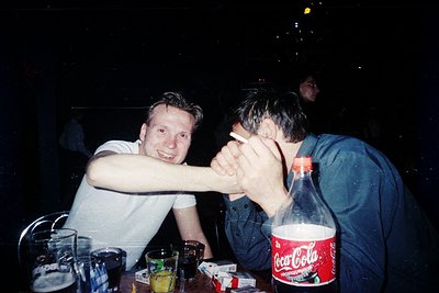 Vintage low-light photo of two men laughing at a dimly lit table, one playfully covering the other’s face. Bottled Coca-Cola ...