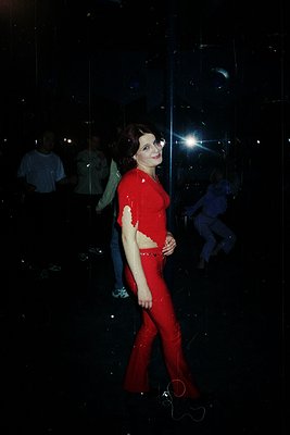A woman in a bold red full-body suit poses in a dimly lit, mirrored disco ball environment, likely from the 1970s–1980s. Refl...