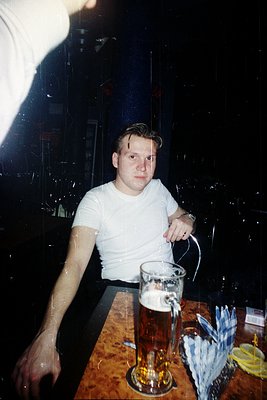 Young man in a sleeveless white shirt at a bar, pouring beer into a tall glass. Reflective glass shelves and bottles in backg...