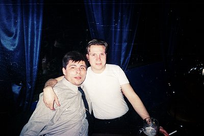 Two men pose indoors under blue stage lighting, likely at a 1990s nightclub or concert. The man on the left wears a light-col...