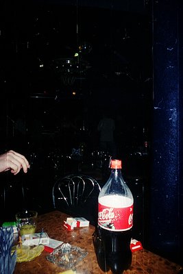Vintage Coca-Cola bottle on a dark wooden table beside a black metal chair, with a glass and scattered wrapping paper. Reflec...