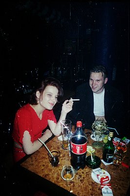 Vintage 1960s diner scene: couple in retro attire (woman in red blouse, man in white shirt) seated at a dark wood table with ...