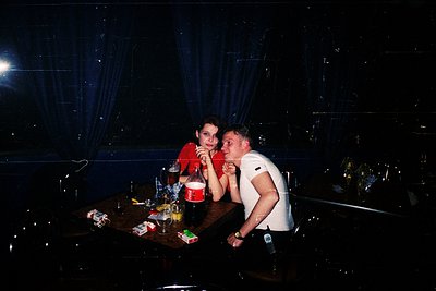 Two individuals share an intimate moment at a dimly lit, modern bar. The woman in red leans in close to the man in white, who...