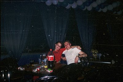 Vintage candid shot of two adults embracing at a dimly lit table, likely a 1970s-1980s social gathering. Bottles of beer, gla...