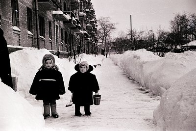 Two children in winter coats and hats walk a snow-covered urban street, one carrying a bucket. Snowbanks line the sidewalk be...